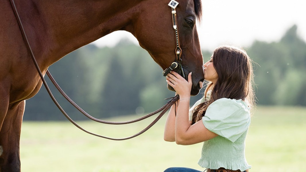 A woman kisses her horse's nose while kneeling in a green pasture in spring.